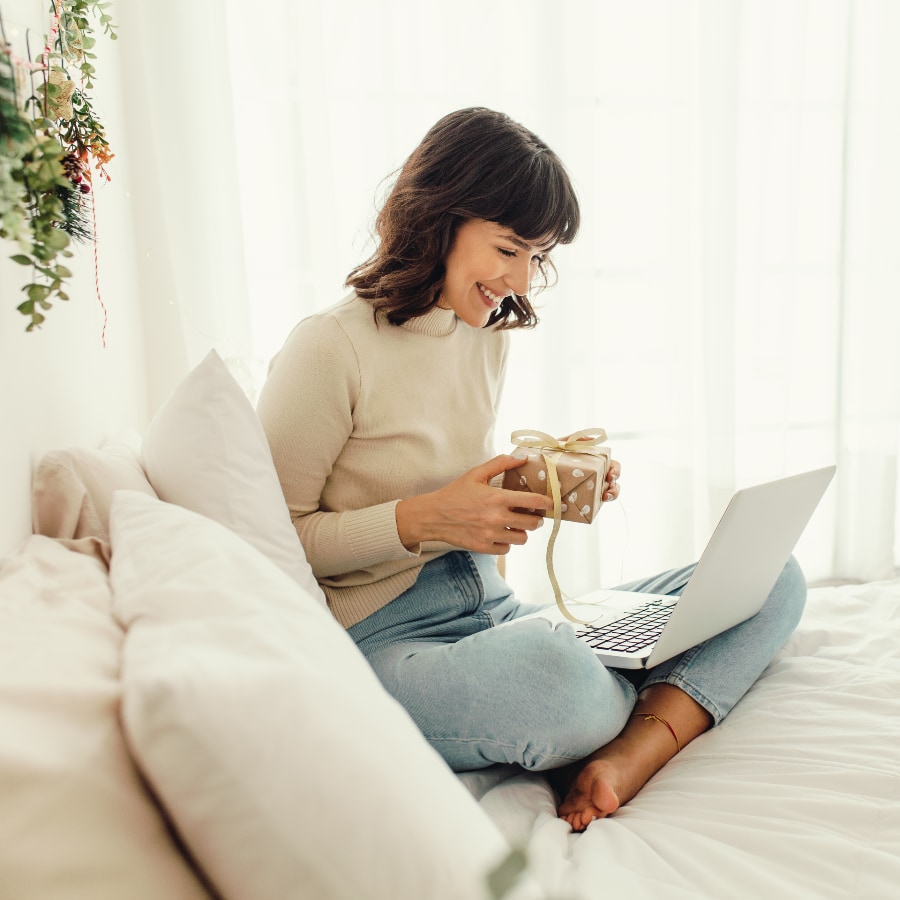 a young brown haired woman sitting on a bed holding a gift while looking at a laptop